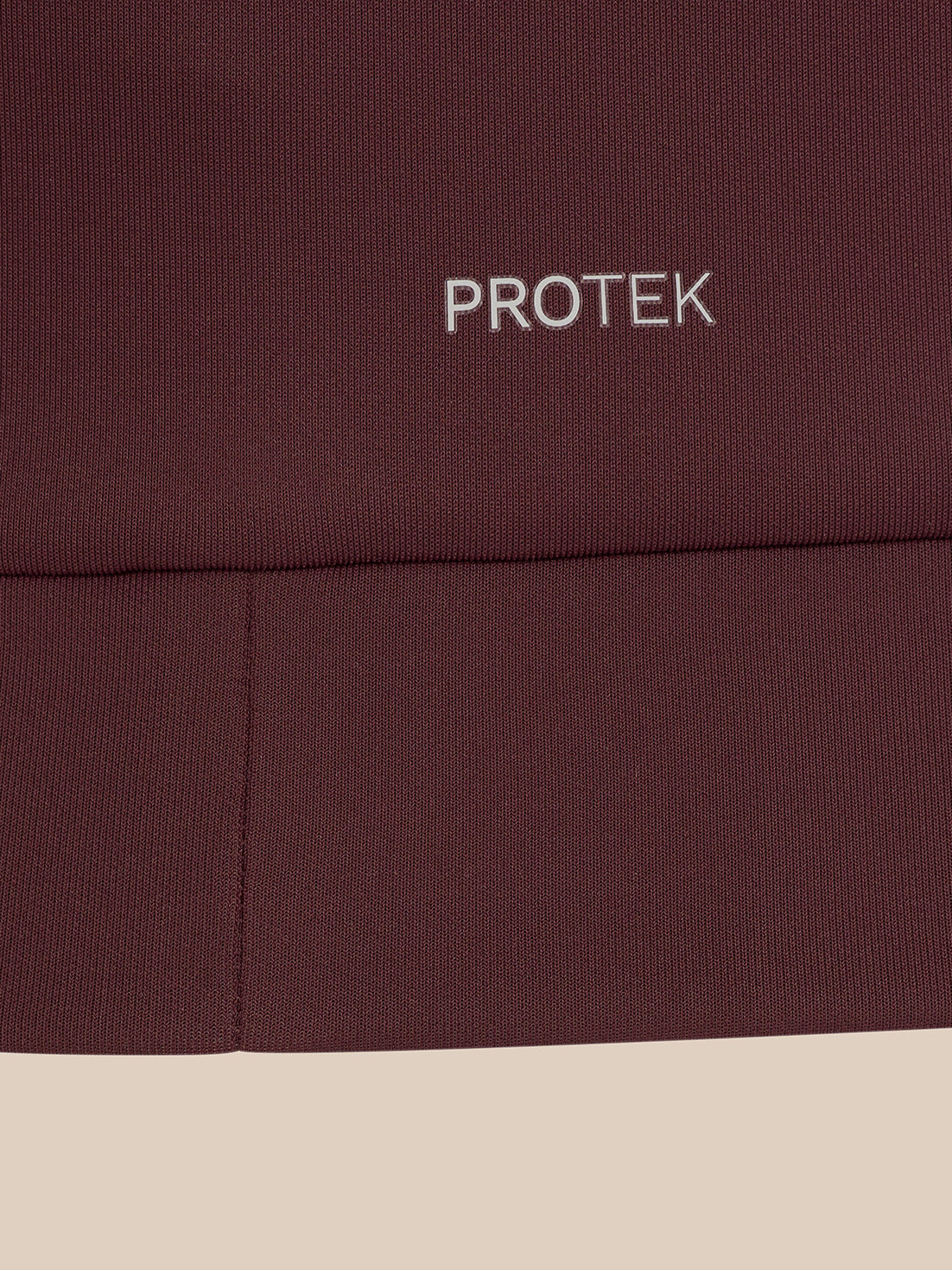 Close-up of a dark burgundy ribbed fabric with the word PROTEK embroidered in white near the top, set against a light beige background.
