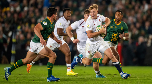 England rugby player in white charges with the ball while green jersey opponents pursue in a crowded stadium.
