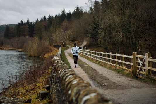 Runner wearing a light gray top jogs along a riverside path with leafless trees and a stone wall.