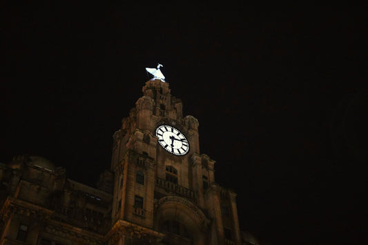 Nighttime view of a clock tower topped by a bright white statue against a dark sky.
