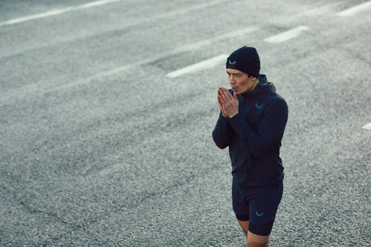 Athlete wearing a navy running jacket and beanie stands on a grey outdoor track with hands pressed together.