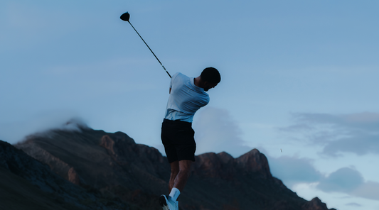 Golfer in a light blue shirt and dark shorts swings a club against a rocky coastal backdrop.