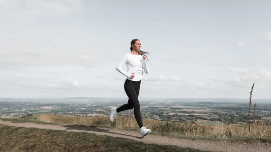 Woman in white long sleeve top and black leggings runs along a hillside trail with a wide rural valley in the background.