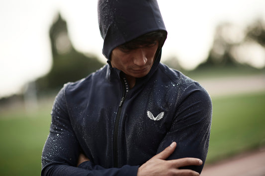 Athlete in a navy rain jacket with a white wing logo stands outdoors in the rain.