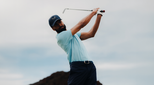 Golfer in a light blue polo and dark cap mid swing against a pale sky.