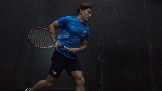 Male tennis player in a blue athletic shirt and dark shorts with a racket on a dark court backdrop.