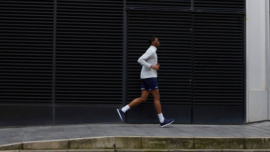 Runner wearing a light jacket and navy shorts jogs along a city sidewalk beside a dark industrial wall.