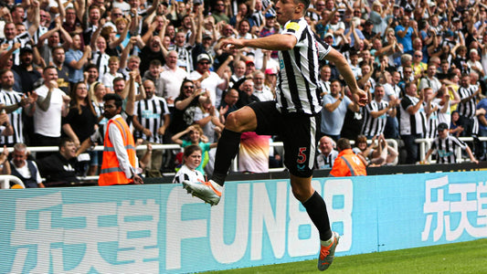 Footballer in black and white striped kit leaps near the sideline as fans celebrate in the stands