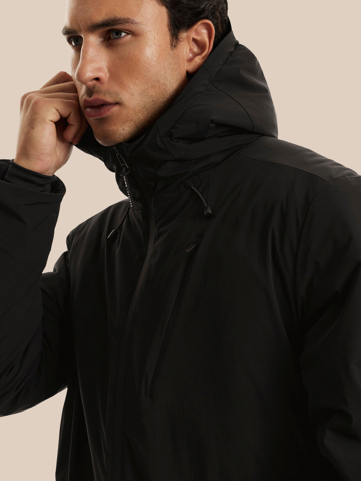 A close-up of a man wearing a black hooded jacket, lifting the collar with his right hand, set against a solid beige background.