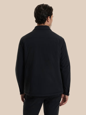 A man viewed from the back wearing a black long-sleeve collared jacket and dark trousers against a solid beige background.