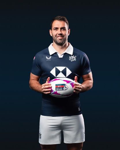 Man holding a rugby ball wearing a dark blue sports jersey with white accents on a black background