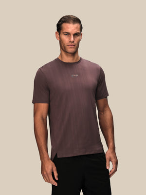 A man wearing a short-sleeve crewneck athletic t-shirt in a muted plum-brown color, paired with black pants, standing against a solid beige background.