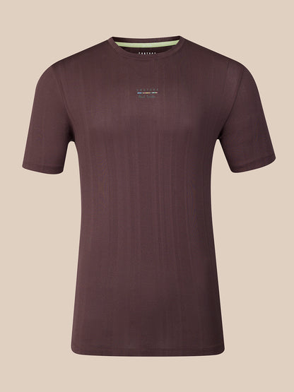 A dark brown short-sleeve athletic t-shirt with a subtle vertical ribbed texture, featuring a small multicolor logo on the center chest and a light green inner collar lining, shown against a solid beige background.