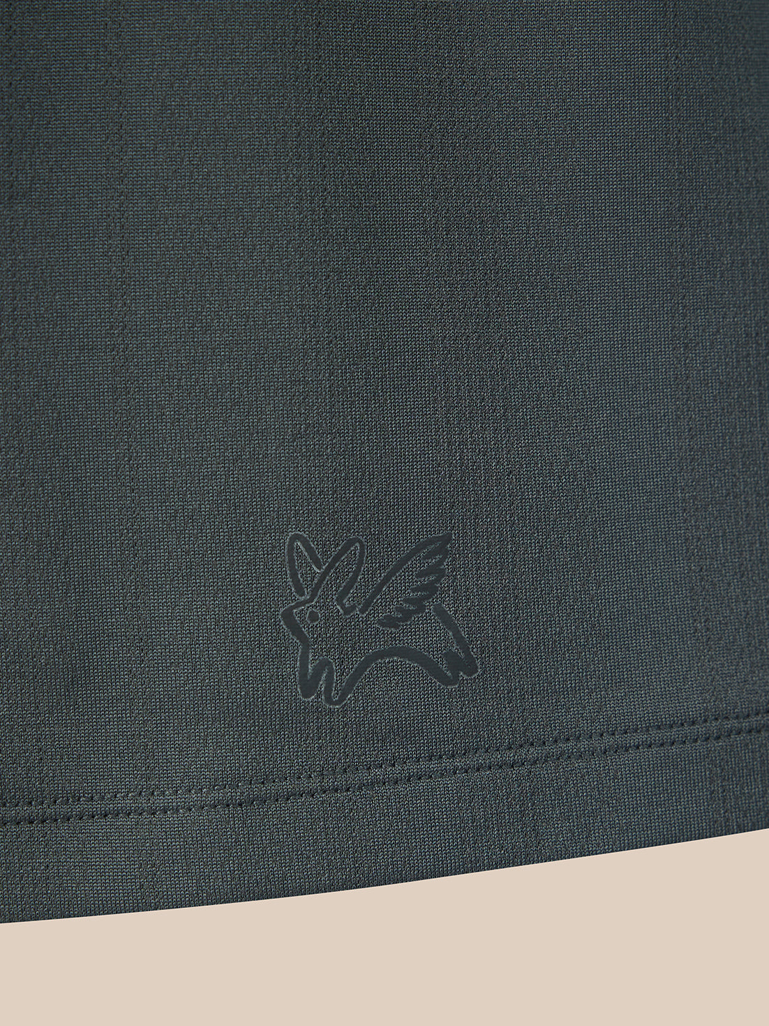 Close-up of a dark charcoal gray knit fabric with a tonal embroidered bird logo near the hem, shown against a light beige background.