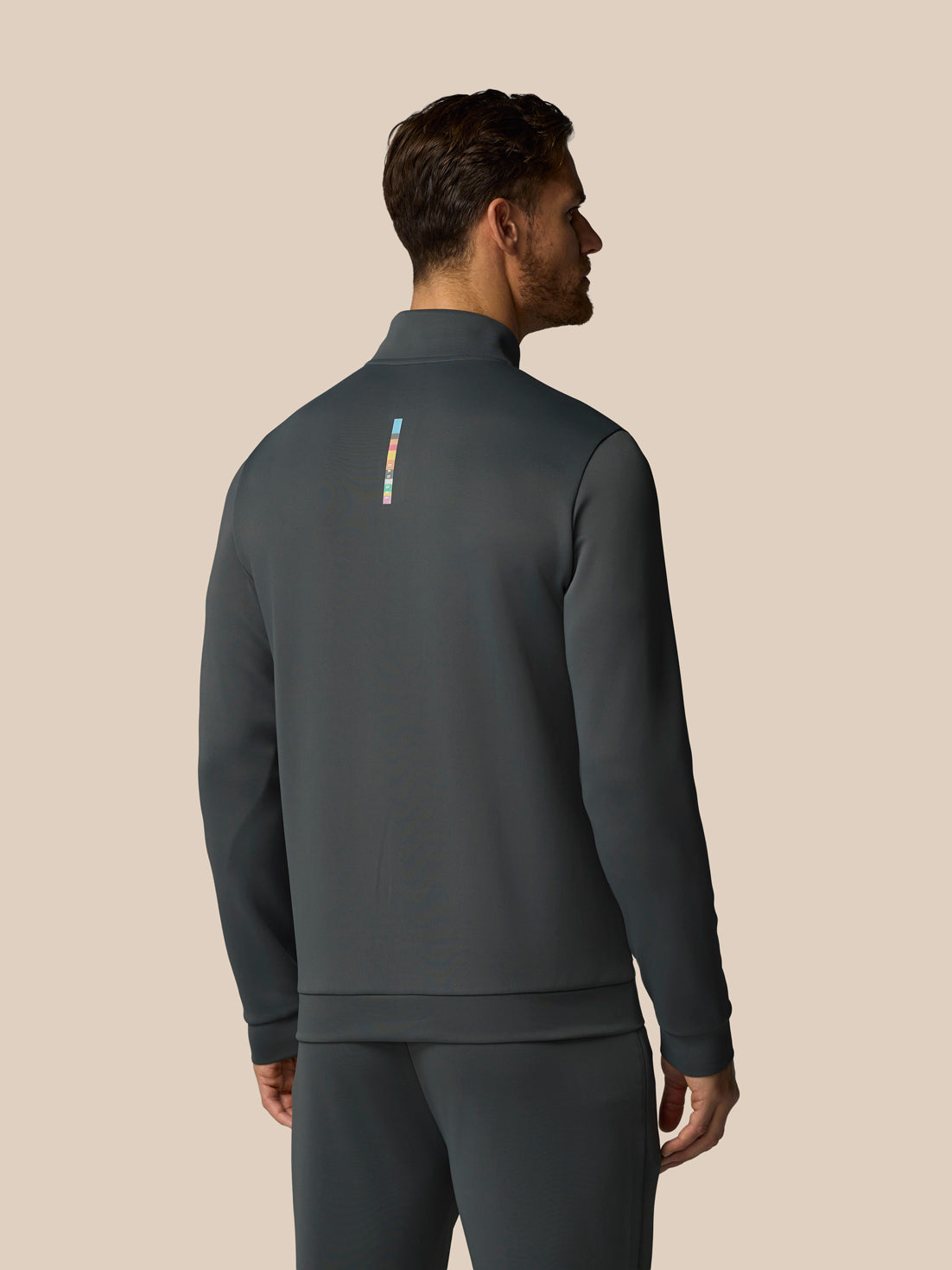 Man wearing a dark gray athletic jacket with a small logo on the back against a beige background
