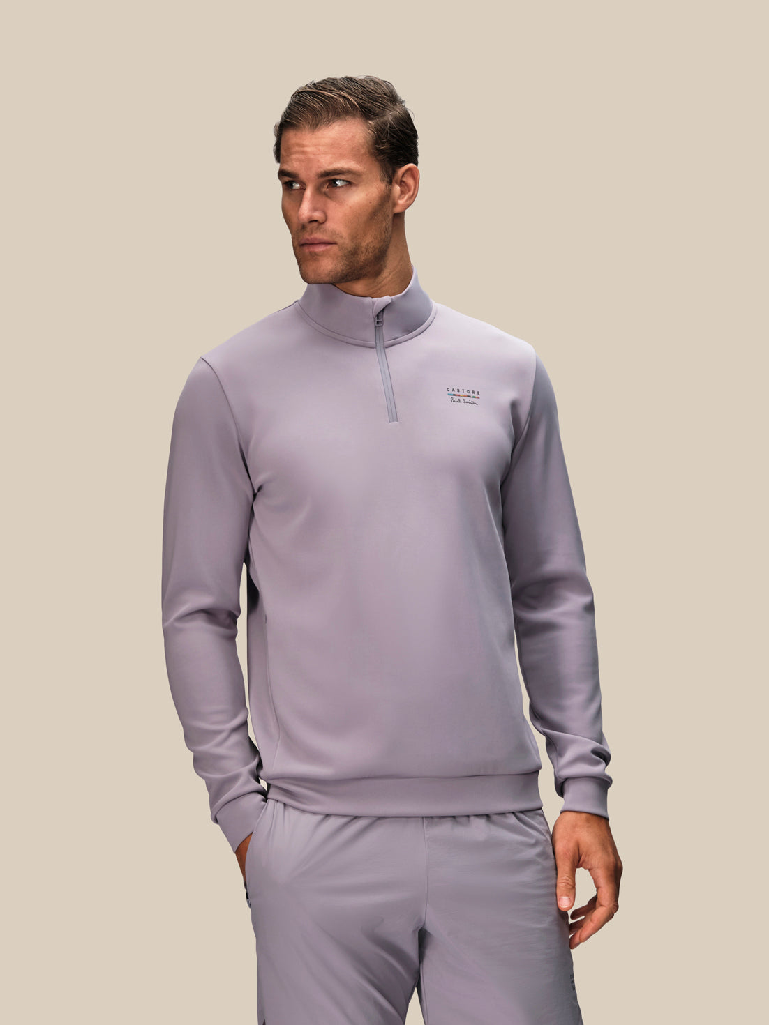 A man modeling a light purple-gray quarter-zip pullover sweatshirt and matching sweatpants against a solid beige background.