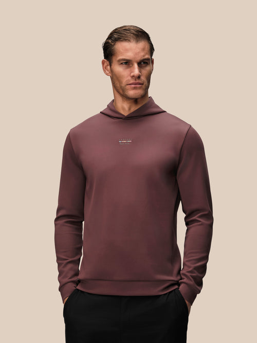 A man wearing a long-sleeve hooded sweatshirt in a dusty rose or mauve color, paired with black pants, standing against a solid beige background.