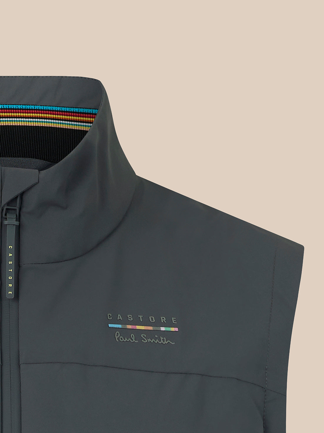 Close-up of a dark gray sleeveless zip-up vest with a high collar, featuring a multicolor stripe detail on the inner collar, a vertical zip tab with 'CASTORE' text, and 'CASTORE Paul Smith' embroidered on the chest, set against a solid beige background.