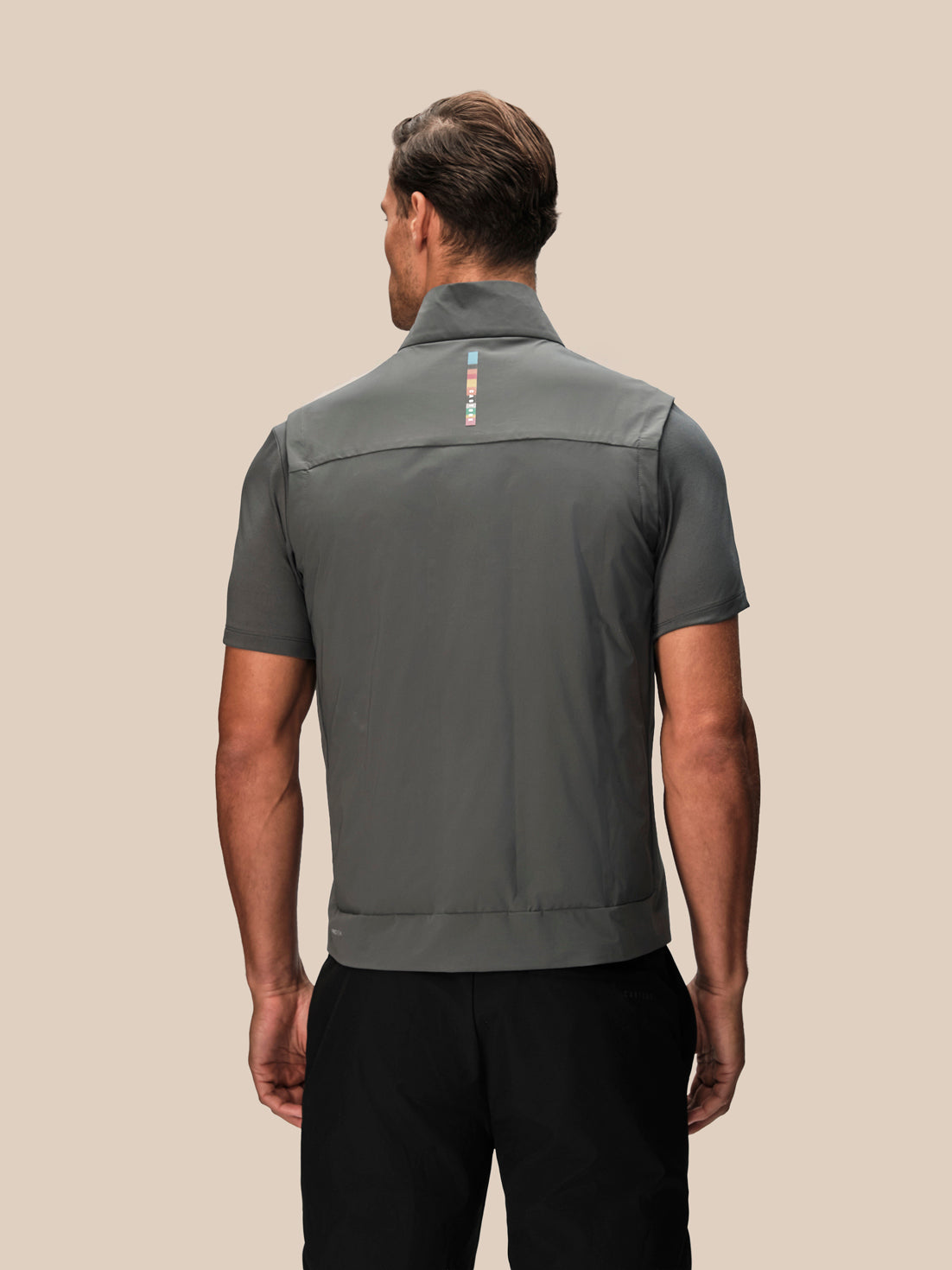 A man viewed from the back wearing a dark gray short-sleeve performance shirt with a small vertical multicolor logo on the upper back, paired with black pants, against a solid beige background.