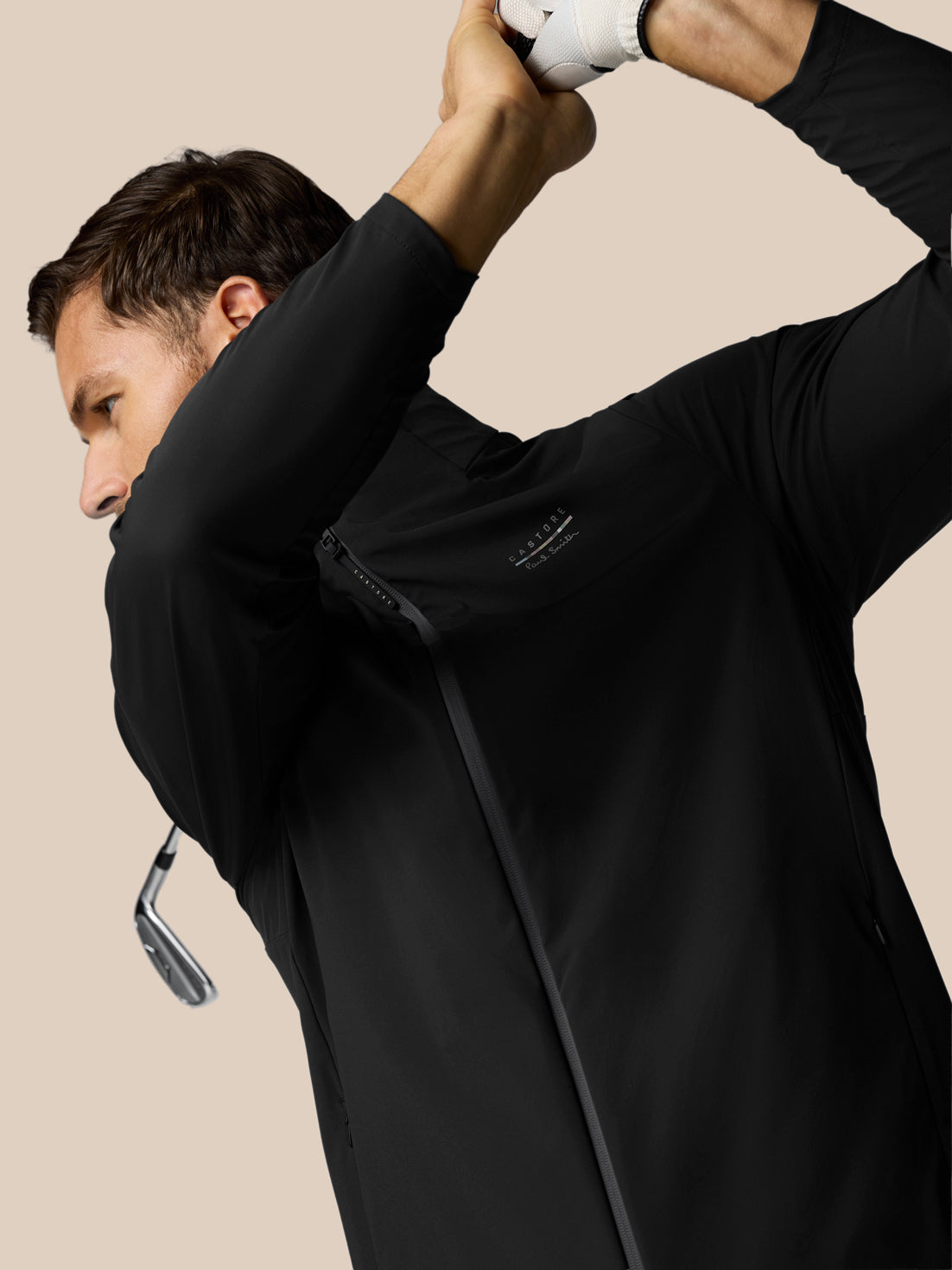 Man in black jacket holding a golf club against a beige background