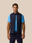 Male model wearing navy Pinnacle Gilet over a light blue polo against a beige studio backdrop