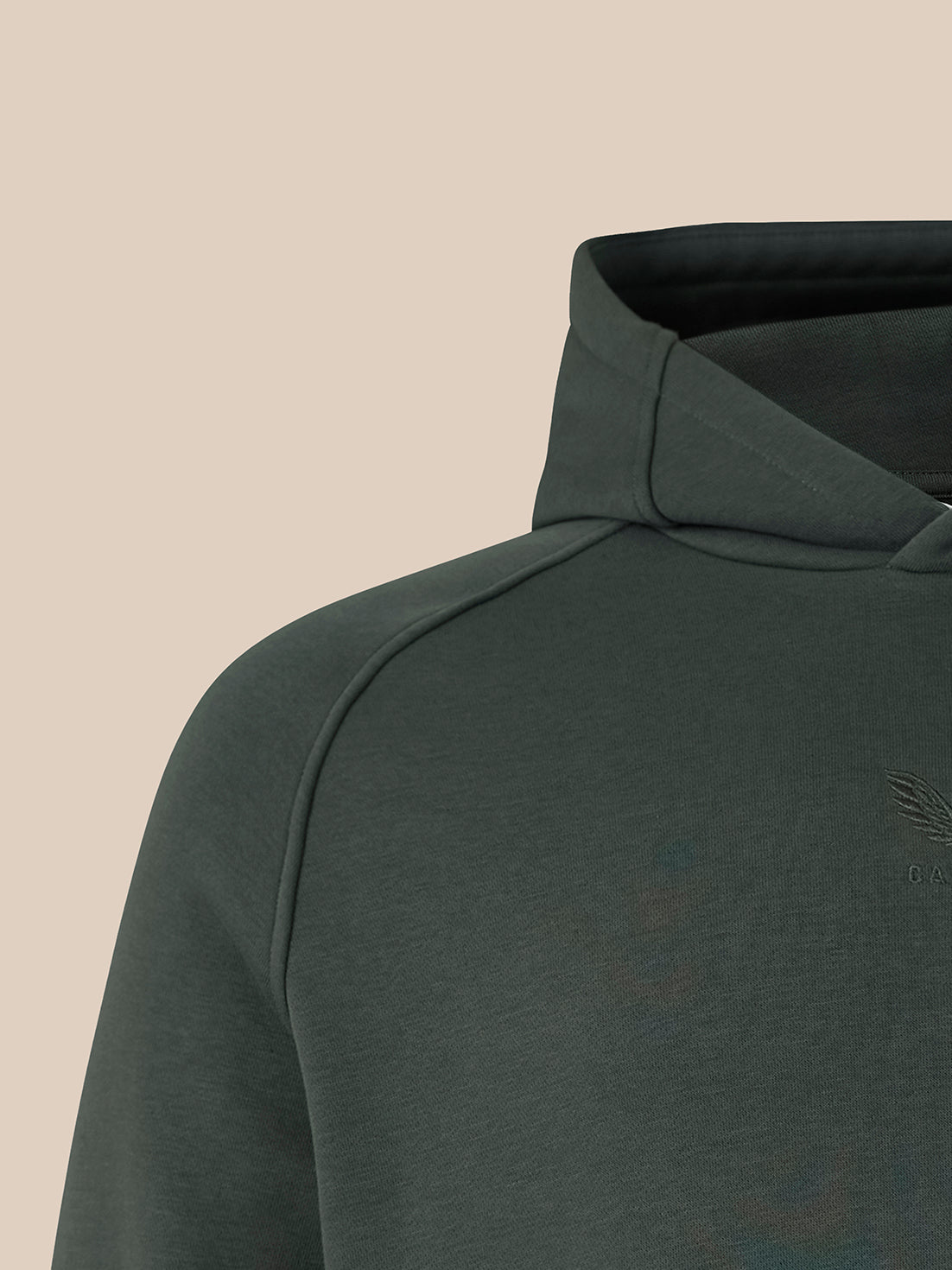 Close-up of a dark gray-green hooded sweatshirt showing the shoulder, collar, and a subtle tonal embroidered chest logo against a solid beige background.