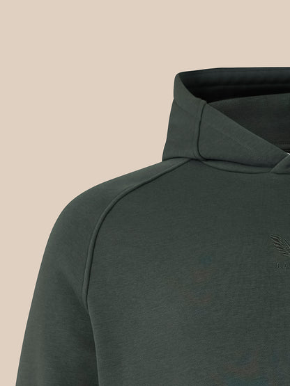 Close-up of a dark gray-green hooded sweatshirt showing the shoulder, collar, and a subtle tonal embroidered chest logo against a solid beige background.