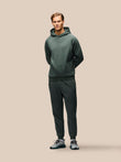 A man modeling a dark green two-piece tracksuit consisting of a pullover hoodie and matching jogger pants, paired with gray and black sneakers, against a solid beige background.