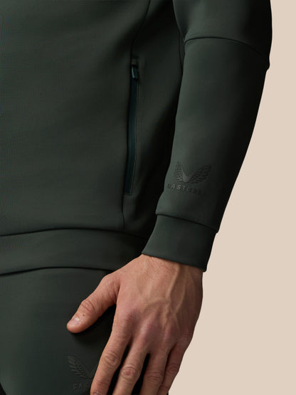 Close-up of a person wearing a dark green CASTORE tracksuit with a beige background
