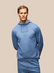 A man wearing a light blue hooded sweatshirt and matching sweatpants against a solid beige background. The hoodie features the brand name 'CASTORE' printed in black on the chest.