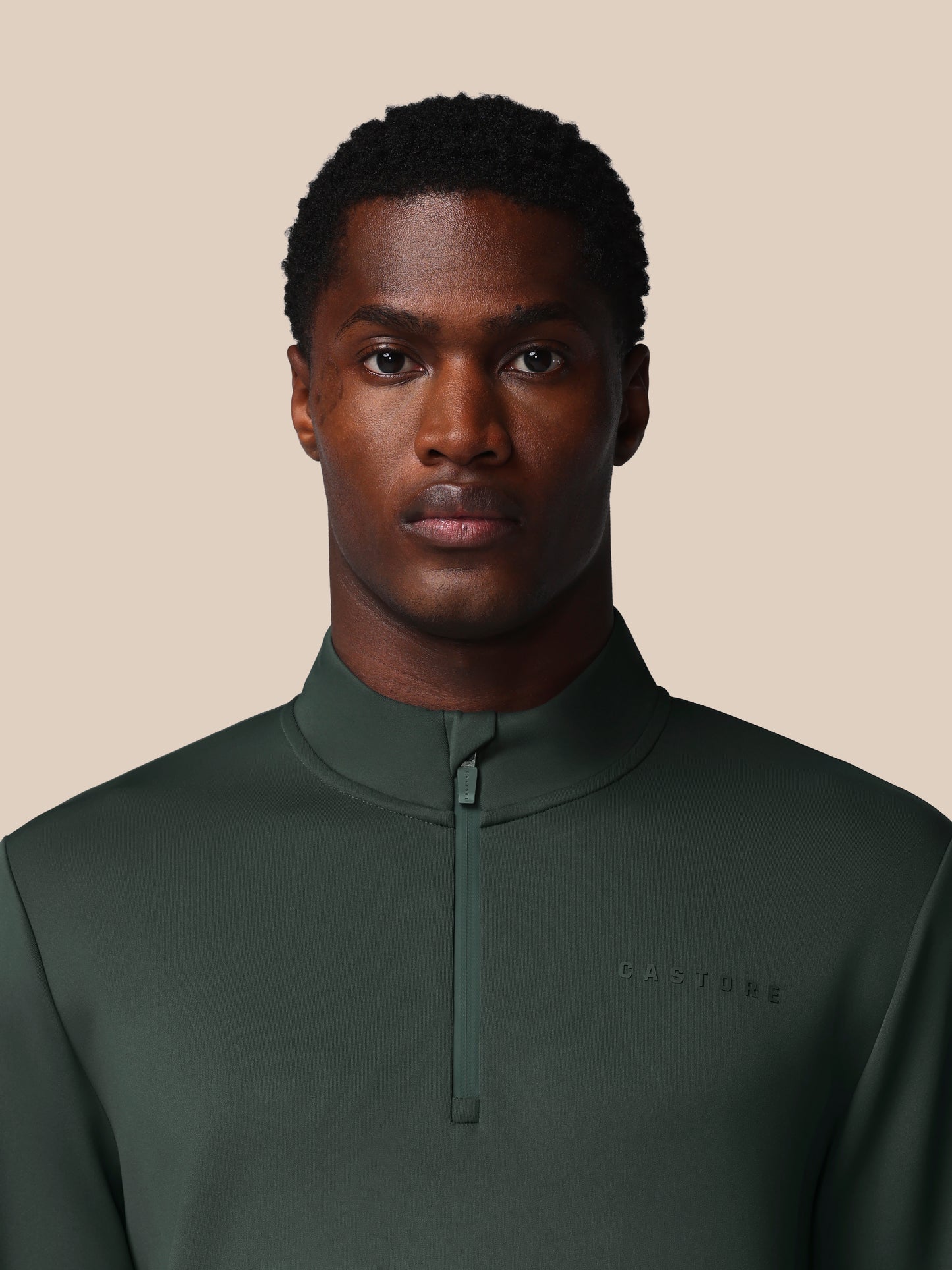 Man wearing a dark green pullover with a brand logo on a beige background