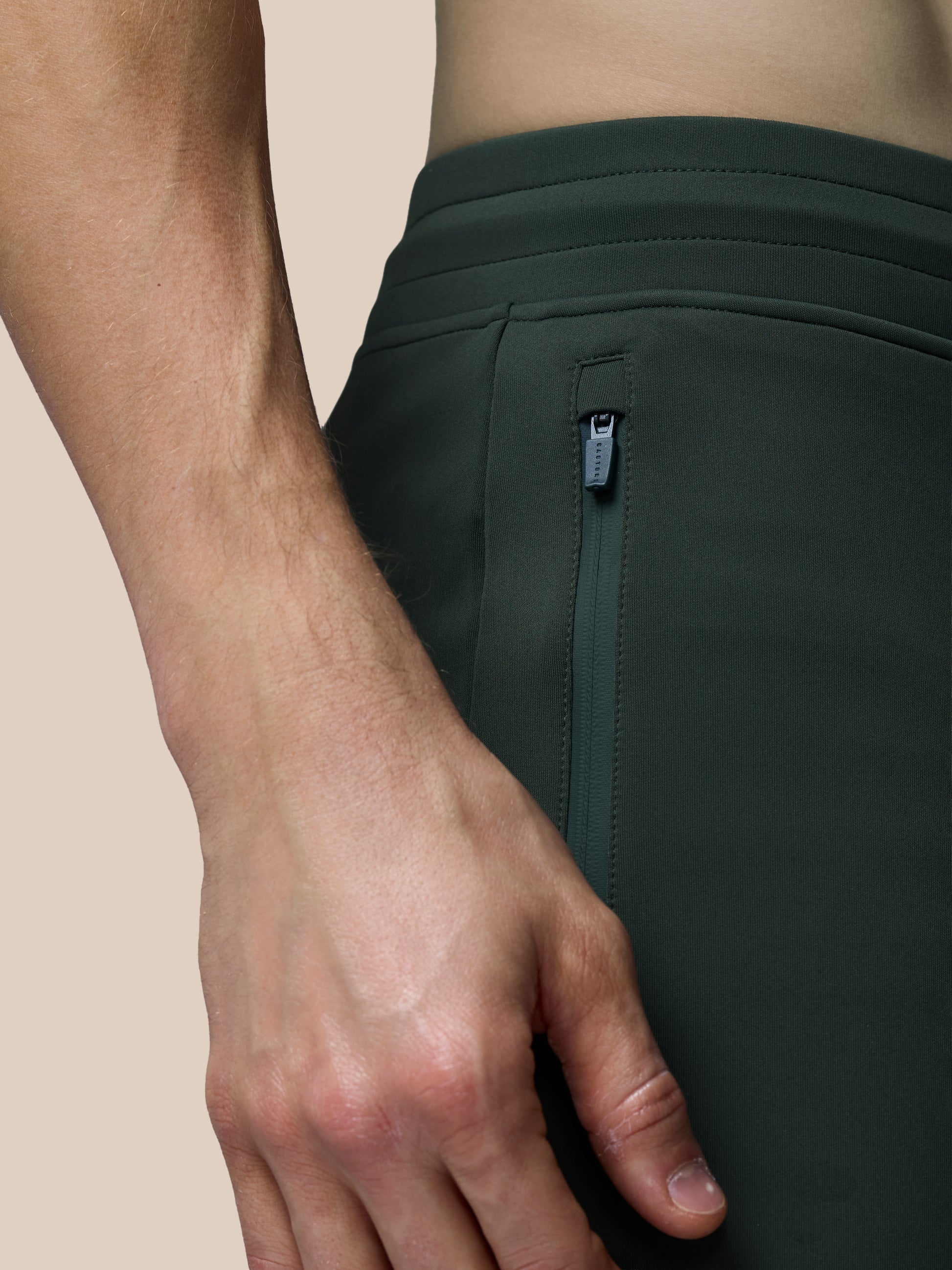 Close-up of a person wearing dark green athletic shorts with a zipper on a beige background