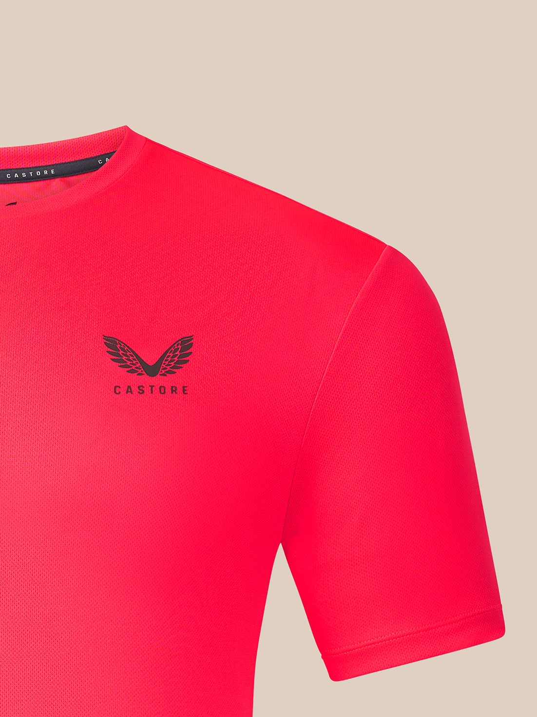 A close-up of a bright pink Castore short-sleeve athletic shirt with a black winged logo and brand name on the left chest, set against a solid beige background.