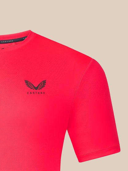 A close-up of a bright pink Castore short-sleeve athletic shirt with a black winged logo and brand name on the left chest, set against a solid beige background.