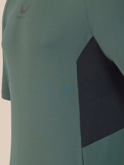 Close-up of a short-sleeve athletic top in muted sage green with a dark charcoal side panel and a small dark logo on the chest.