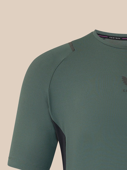 Close-up of a short-sleeve athletic top in muted sage green with a subtle waffle texture, dark side paneling, and a small chest logo, shown against a plain warm beige background.