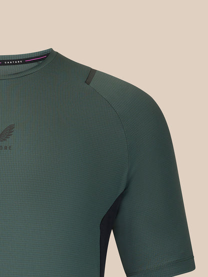 Close-up of a textured short-sleeve athletic top in dark green, showing the chest, shoulder, and collar area against a plain beige background.