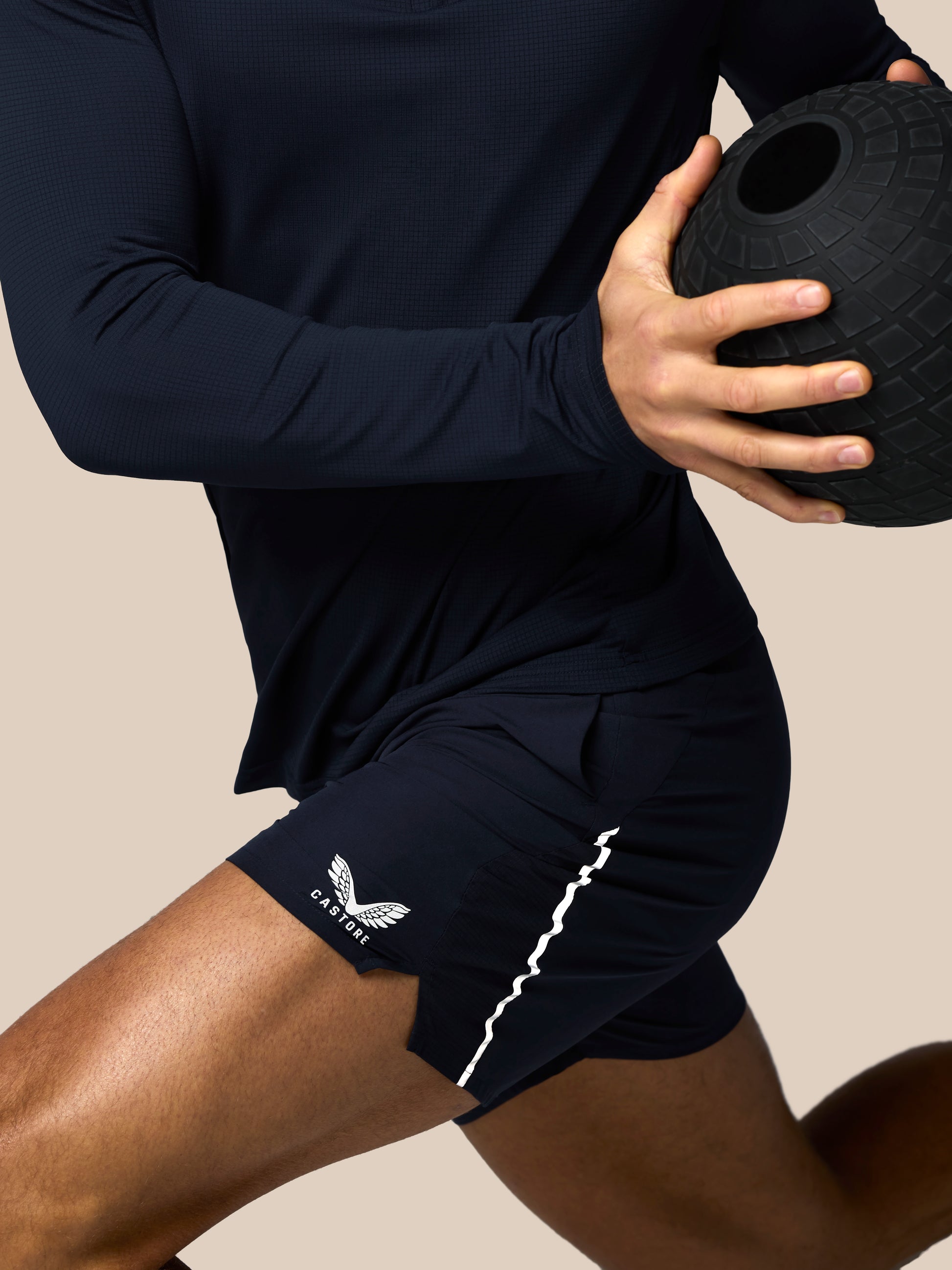 Person wearing navy athletic outfit with a logo, holding a black medicine ball on a beige background