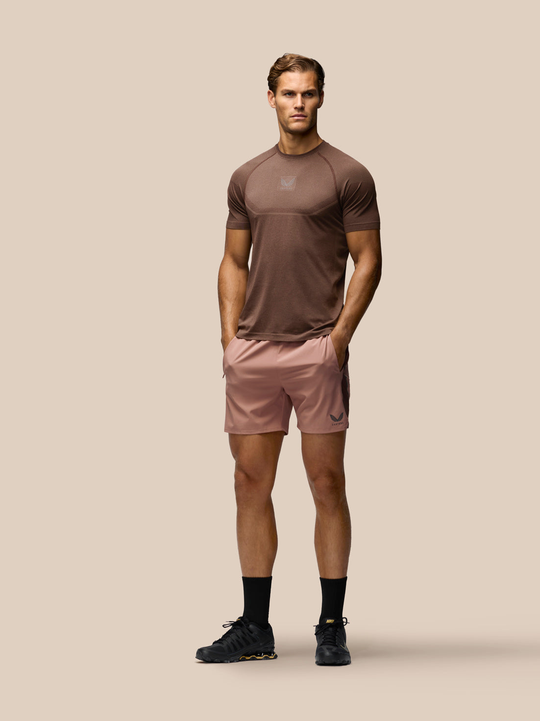 A male model stands against a solid beige background wearing a brown athletic t-shirt, dusty pink athletic shorts, black socks, and black running shoes.