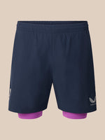 A pair of dark navy blue athletic shorts with a built-in purple inner layer visible at the hem, featuring a white wing logo and the brand name 'CASTORE' on the lower right leg, set against a solid beige background.