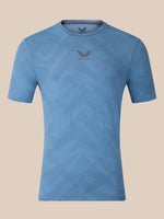 A light blue short-sleeve athletic t-shirt with a subtle geometric pattern and a dark gray wing logo with 'CASTORE' text on the center chest, displayed against a solid beige background.