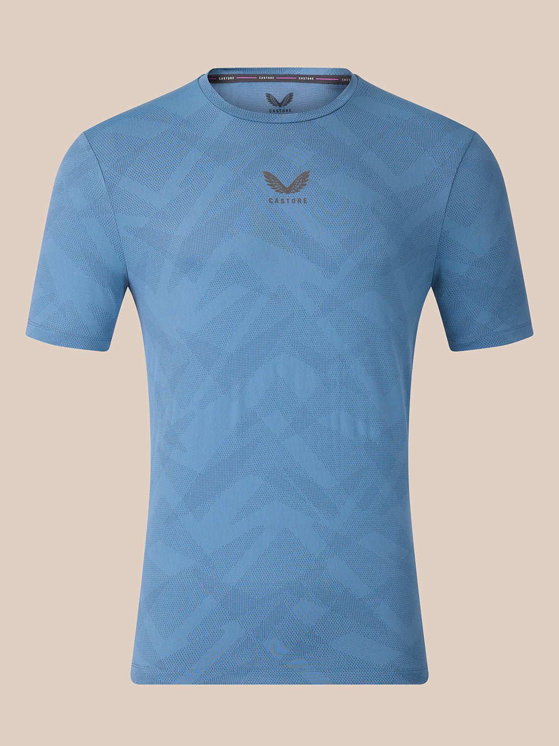 A light blue short-sleeve athletic t-shirt with a subtle geometric pattern and a dark gray wing logo with 'CASTORE' text on the center chest, displayed against a solid beige background.