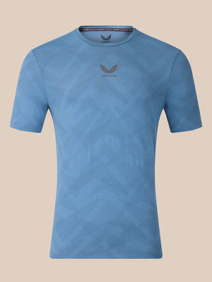 A light blue short-sleeve athletic t-shirt with a subtle geometric pattern and a dark gray wing logo with 'CASTORE' text on the center chest, displayed against a solid beige background.