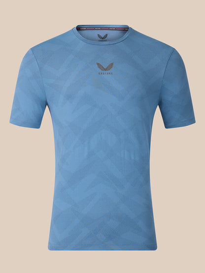 A light blue short-sleeve athletic t-shirt with a subtle geometric pattern and a dark gray wing logo with 'CASTORE' text on the center chest, displayed against a solid beige background.