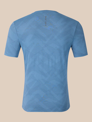 A light blue short-sleeve t-shirt with a subtle geometric pattern, shown from the back against a solid beige background. The brand name 'CASTORE' is printed vertically near the upper back neckline.