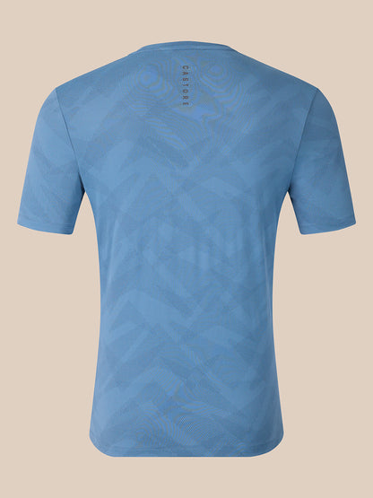 A light blue short-sleeve t-shirt with a subtle geometric pattern, shown from the back against a solid beige background. The brand name 'CASTORE' is printed vertically near the upper back neckline.