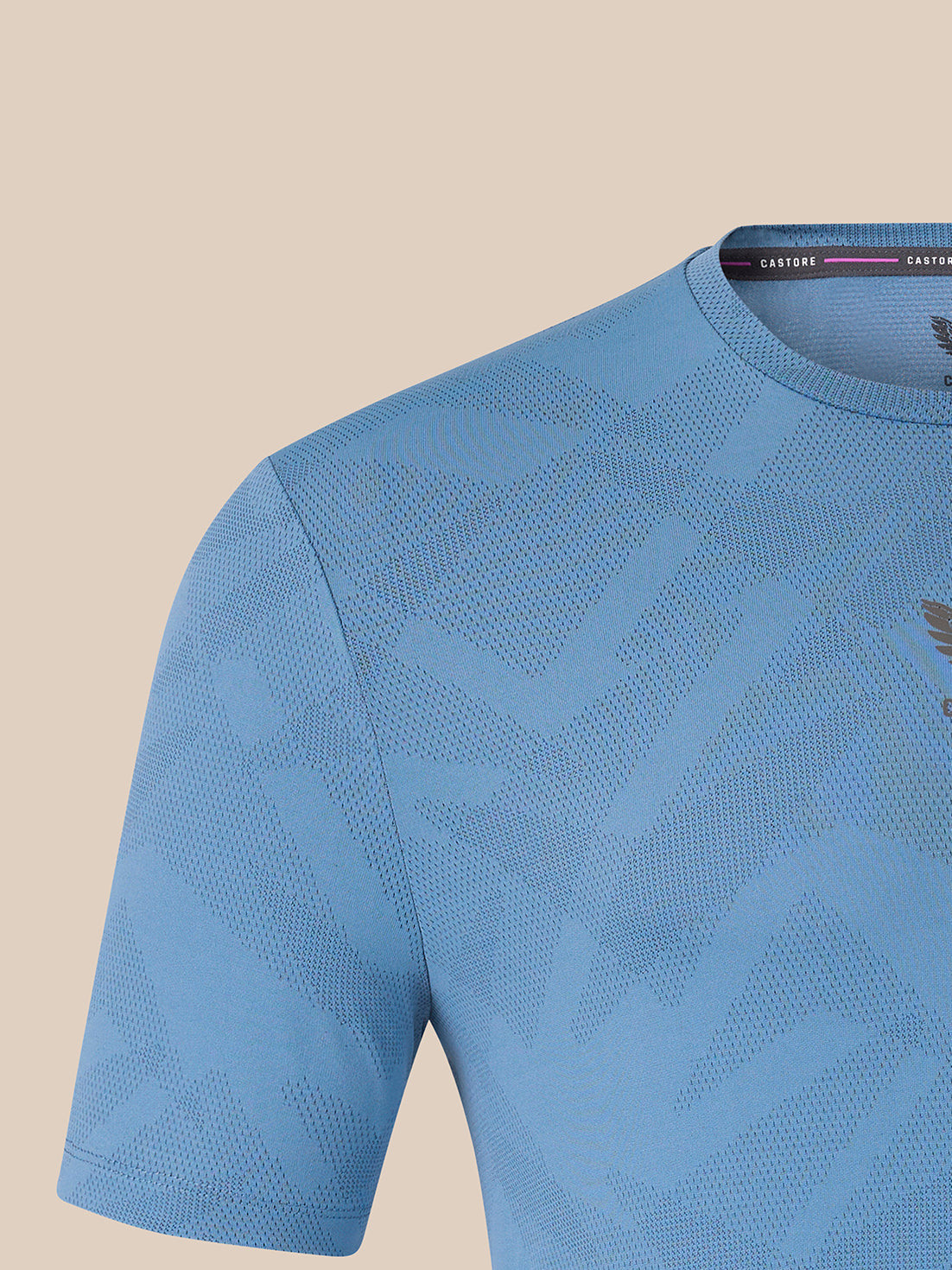 A close-up of a light blue short-sleeve athletic shirt with a subtle geometric pattern and a small dark logo on the chest, set against a solid beige background.