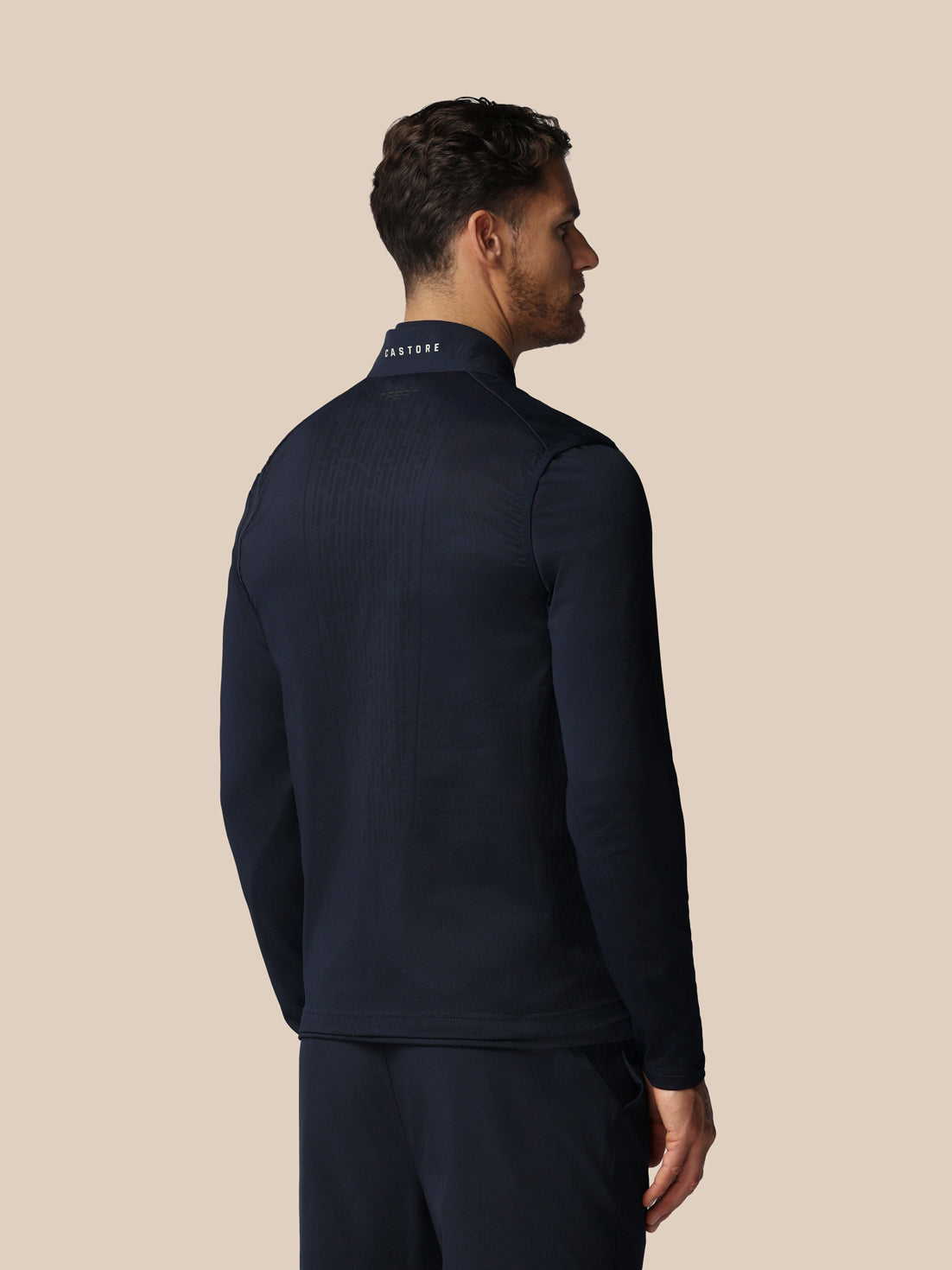 Man wearing a navy blue jacket with 'Castore' branding on the back against a beige background