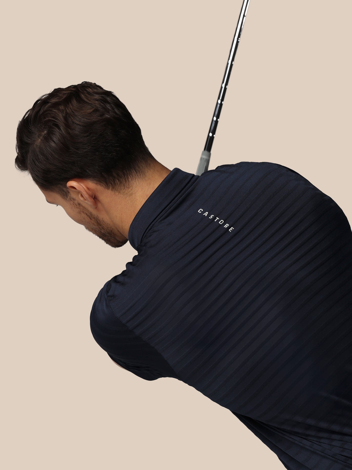 Man in a navy blue shirt with 'castore' branding holding a golf club on a beige background