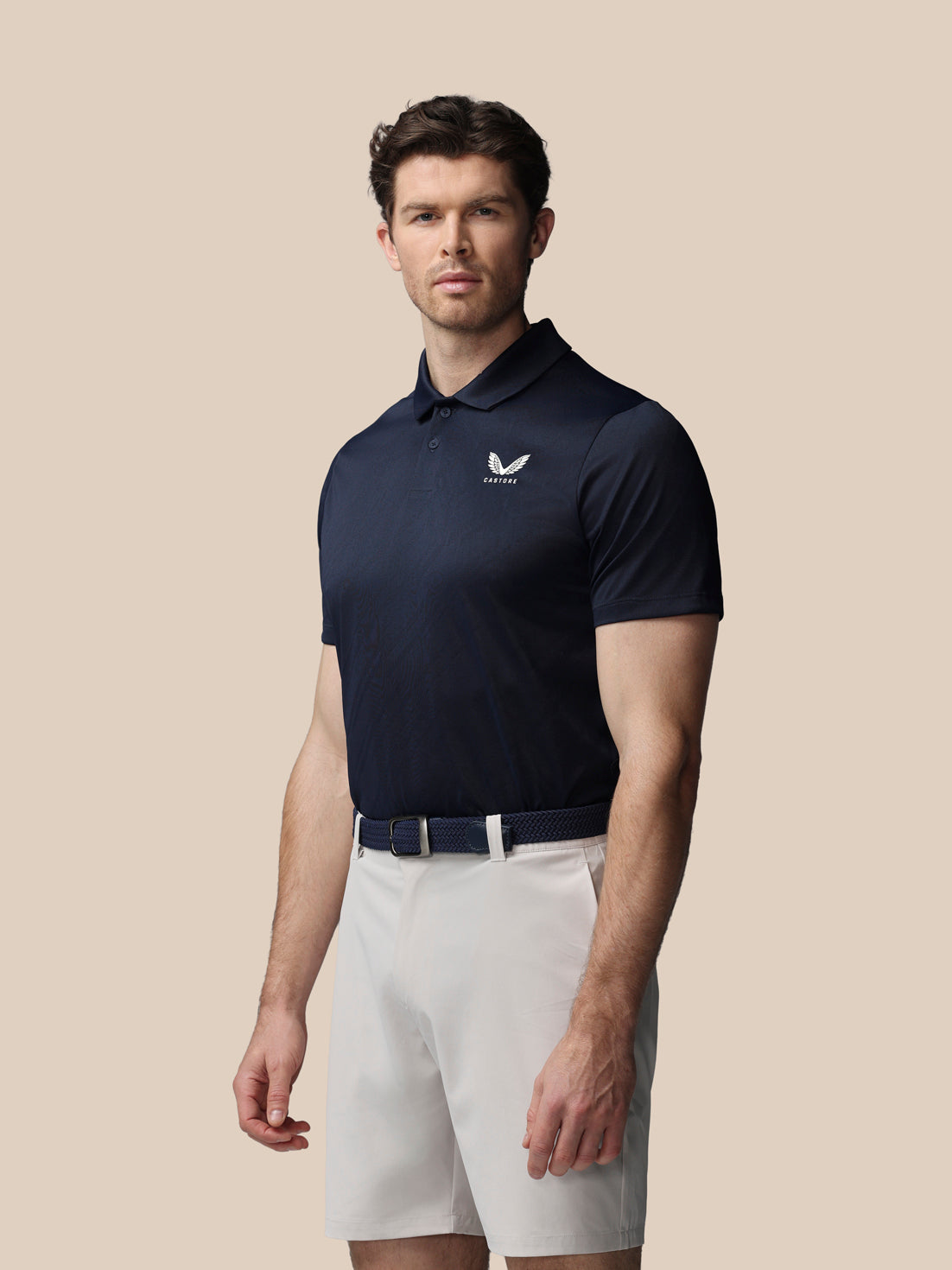 Man wearing a navy polo shirt with a logo and white shorts on a beige background